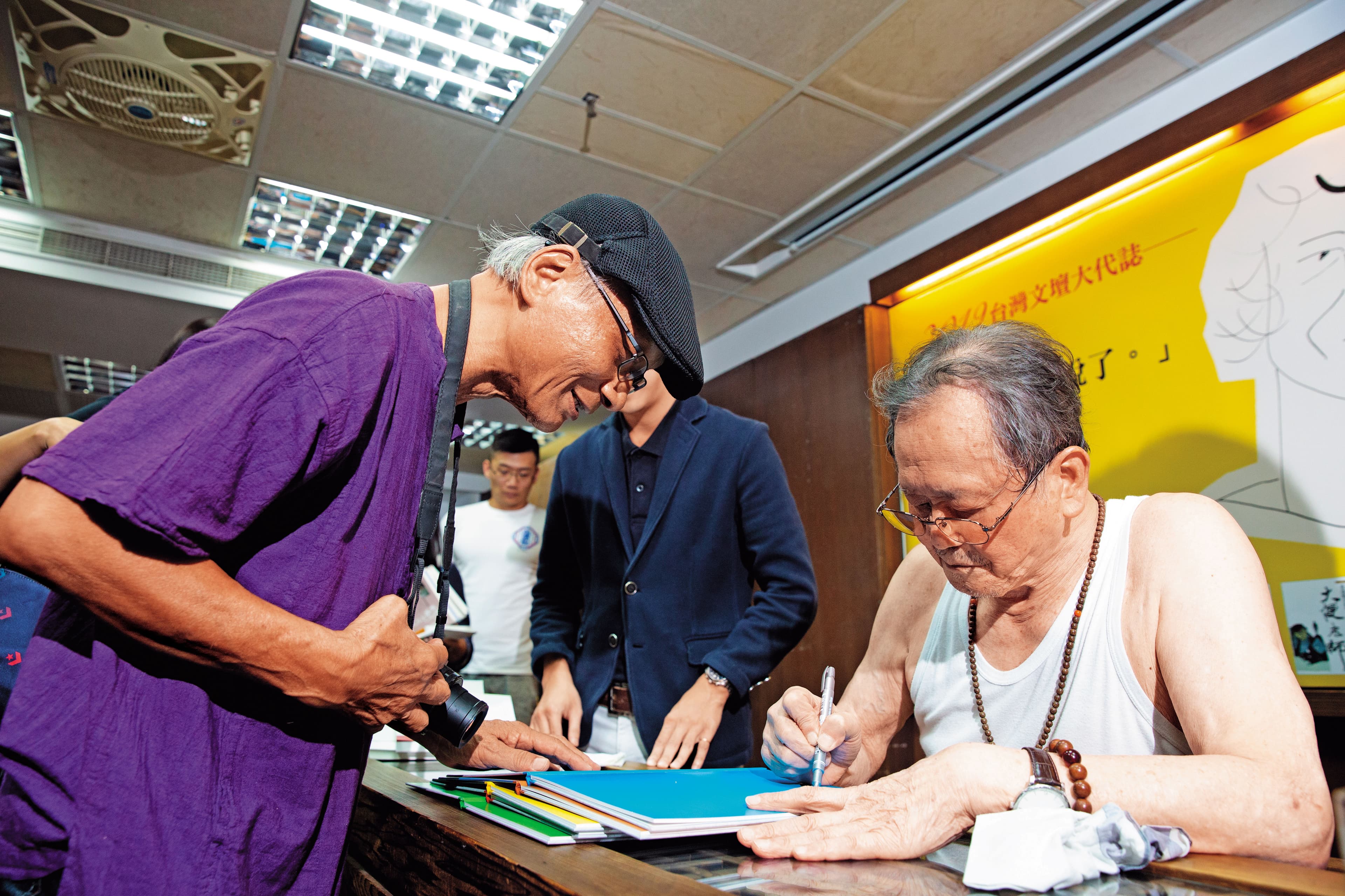 黃春明（右）新書座談會簽得滿頭大汗，索性扒下襯衫，就穿著一件背心振筆疾書。