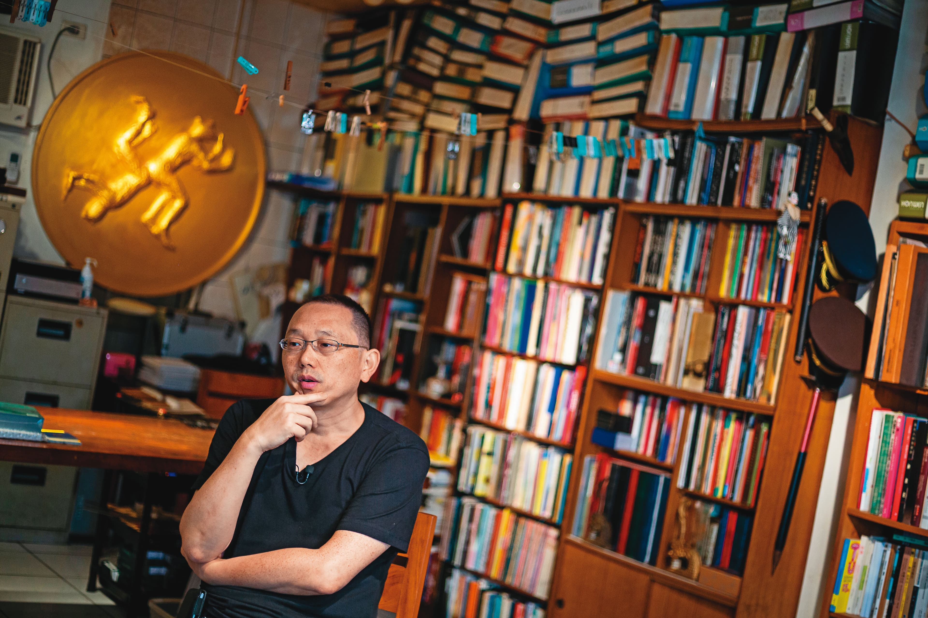 姚瑞中工作室(幻影堂)收藏了許多藝術書籍與展冊文獻,他身後的金色圓徽是他為今年大型個展《犬儒共和國》設計的國徽。
